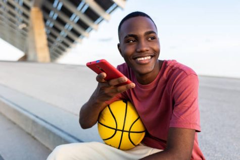 Happy young black man with basquetball ball relaxing outdoors while using smart mobile phone to send an audio voice message - Technology, youth and millennial generation people concept
